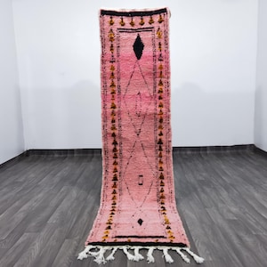 Op de afbeelding: Een roze en zwart loper met een geometrisch ontwerp. Het tapijt heeft een centraal ruitpatroon en decoratieve randen met kwastjes aan de onderkant. Het tapijt is gemaakt van geweven materiaal.