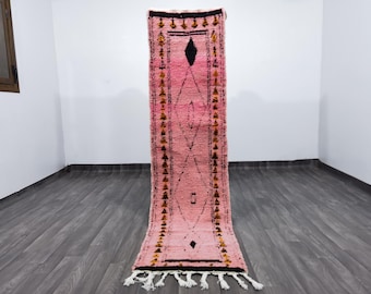 May include: A pink and black runner rug with a geometric design. The rug features a central diamond pattern and decorative borders with tassels at the bottom. The rug is made of woven material.