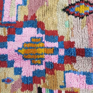 May include: Close-up of a colorful, textured rug with a geometric pattern. The rug features a mix of pink, blue, red, yellow, and brown hues. The design includes squares, rectangles, and diamond shapes, creating a vibrant and eye-catching textile.