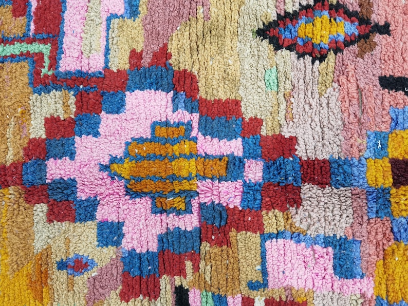 May include: Close-up of a colorful, textured rug with a geometric pattern. The rug features a mix of pink, blue, red, yellow, and brown hues. The design includes squares, rectangles, and diamond shapes, creating a vibrant and eye-catching textile.