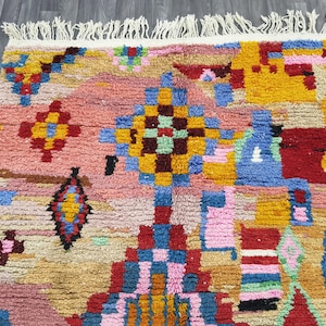 May include: A colorful, patterned rug with a textured surface. The rug features a mix of geometric shapes and designs in shades of pink, yellow, blue, red, and green. The rug has a fringed edge and is likely made of wool or a similar material.