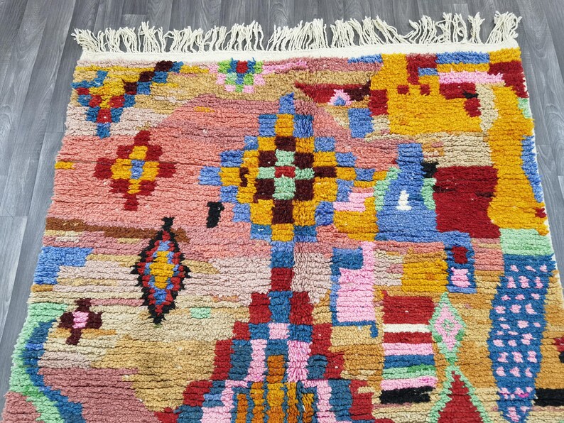 May include: A colorful, patterned rug with a textured surface. The rug features a mix of geometric shapes and designs in shades of pink, yellow, blue, red, and green. The rug has a fringed edge and is likely made of wool or a similar material.