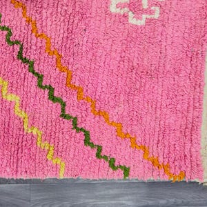 May include: A bright pink area rug featuring a series of diagonal, wavy lines in yellow, dark green, and orange. The rug has a thick, textured pile and is finished with a cream-colored fringe along the right edge, showcasing a geometric pattern.