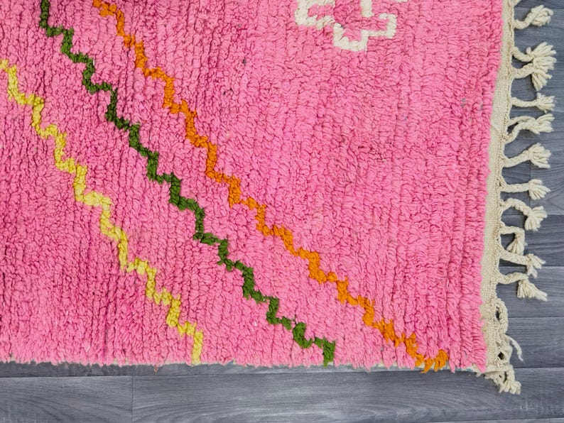 May include: A bright pink area rug featuring a series of diagonal, wavy lines in yellow, dark green, and orange. The rug has a thick, textured pile and is finished with a cream-colored fringe along the right edge, showcasing a geometric pattern.