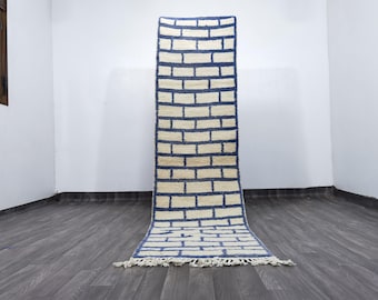May include: A rectangular rug with a brick-like pattern. The rug features a cream-colored background with navy blue rectangular shapes. The rug has a fringe at the bottom and is displayed against a white wall.