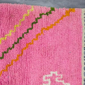 May include: A pink shag rug featuring diagonal zigzag lines in yellow, green, and orange. The rug has a thick, textured pile and is finished with cream-colored fringe tassels along one edge, creating a distinct pattern across the surface.