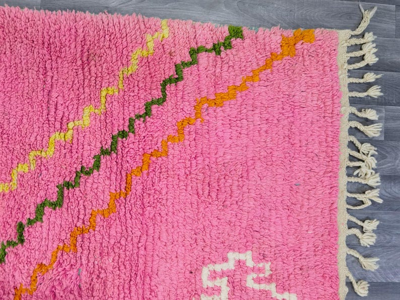 May include: A pink shag rug featuring diagonal zigzag lines in yellow, green, and orange. The rug has a thick, textured pile and is finished with cream-colored fringe tassels along one edge, creating a distinct pattern across the surface.