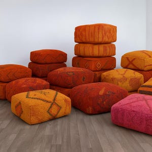 May include: A collection of square, textured floor cushions in shades of orange, red, and pink. The cushions feature geometric patterns and varying textures, creating a warm and inviting aesthetic. Some are stacked, showcasing their size and design.