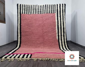 May include: A large, rectangular rug with a pink center and black and white striped borders. A hot pink line accents the bottom. The rug has a textured appearance and a fringe along the bottom edge. The rug is displayed on a dark wood floor.