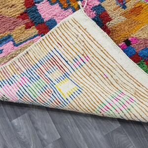 May include: A colorful, hand-woven rug with a geometric pattern. The rug features a mix of pink, blue, green, and orange hues. The underside of the rug is visible, showing a cream-colored backing with vertical stripes.