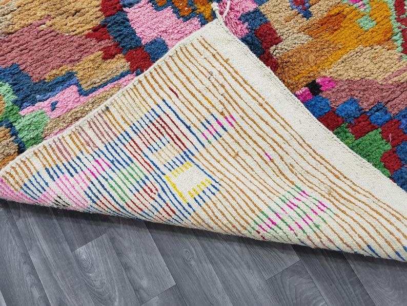 May include: A colorful, hand-woven rug with a geometric pattern. The rug features a mix of pink, blue, green, and orange hues. The underside of the rug is visible, showing a cream-colored backing with vertical stripes.