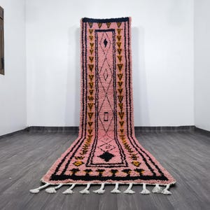May include: A pink and black Moroccan rug with geometric patterns. The rug has a central diamond design with black and yellow accents. White tassels are at the bottom edge. The rug is on a dark grey wooden floor.