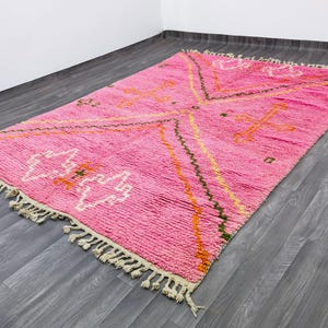 May include: A rectangular area rug featuring a vibrant pink base with geometric patterns in white, orange, and dark green. The rug has a thick, textured pile and is finished with cream-colored knotted fringe tassels along both shorter ends.