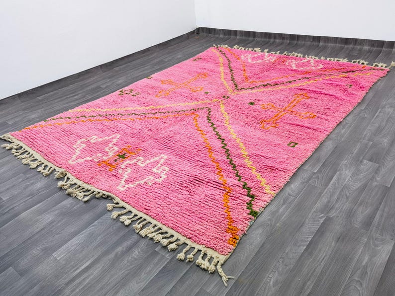 May include: A rectangular area rug featuring a vibrant pink base with geometric patterns in white, orange, and dark green. The rug has a thick, textured pile and is finished with cream-colored knotted fringe tassels along both shorter ends.