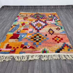 May include: A colorful, rectangular rug with a geometric pattern. The rug features a mix of orange, pink, blue, and green hues. It has a fringe along the bottom edge and is placed on a wooden floor.
