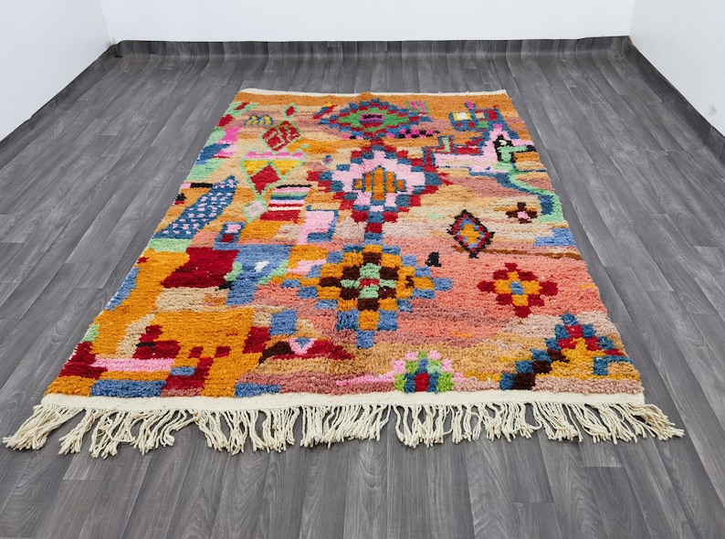 May include: A colorful, rectangular rug with a geometric pattern. The rug features a mix of orange, pink, blue, and green hues. It has a fringe along the bottom edge and is placed on a wooden floor.