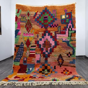 May include: A vibrant, hand-woven rug with a Berber design. The rug features a complex geometric pattern in shades of orange, pink, blue, green, and red. The rug has a textured surface and a fringed edge, adding to its unique appeal.