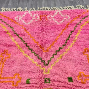 May include: A bright pink area rug featuring a high-pile texture and cream-colored fringe along the top edge. The rug displays geometric patterns in orange, yellow, and dark green, including large cross-like shapes and zigzag lines across the surface.