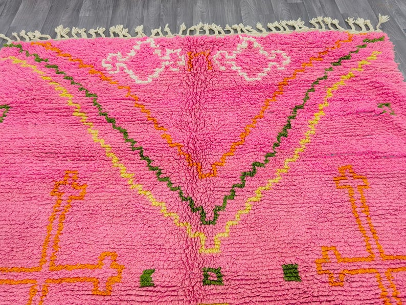 May include: A bright pink area rug featuring a high-pile texture and cream-colored fringe along the top edge. The rug displays geometric patterns in orange, yellow, and dark green, including large cross-like shapes and zigzag lines across the surface.