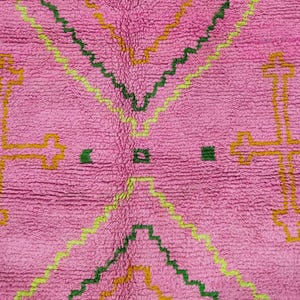 May include: A vibrant pink rug featuring a textured pile surface with geometric patterns. The design includes large yellow cross motifs on the sides and nested zigzag lines in shades of lime green, dark green, and mustard yellow throughout.
