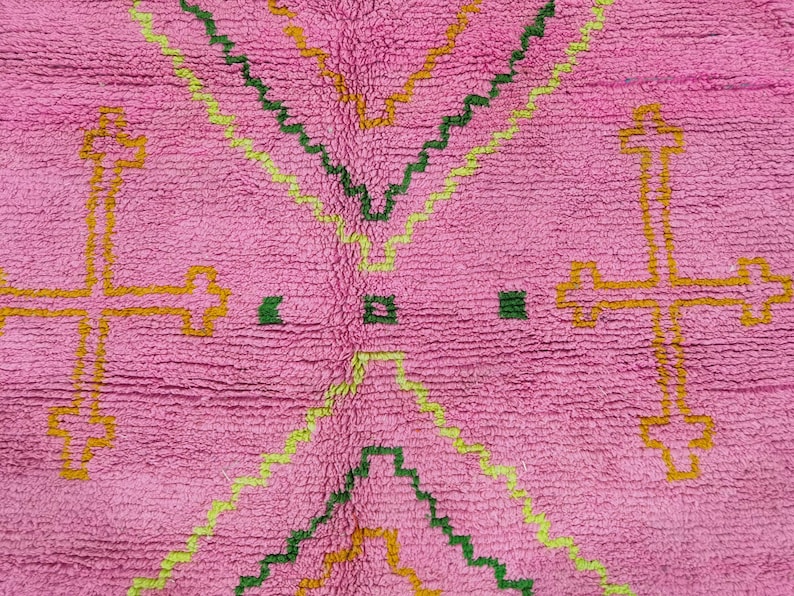 May include: A vibrant pink rug featuring a textured pile surface with geometric patterns. The design includes large yellow cross motifs on the sides and nested zigzag lines in shades of lime green, dark green, and mustard yellow throughout.