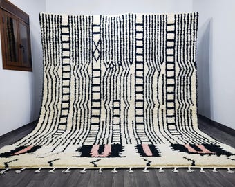 May include: A large, cream-colored rug with a bold black and pink geometric pattern. The design features vertical stripes, ladder-like shapes, and rectangular blocks. The rug has a textured appearance and a fringe along the bottom edge. It is displayed against a white wall.