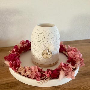 Könnte beinhalten: Eine weiße Keramikvase mit Punktmuster, die auf einem weißen runden Tablett steht. Das Tablett ist von einem Kranz aus rosa und roten Trockenblumen umgeben. Ein kleines Etikett mit Text und einem Herz ist an der Vase befestigt.