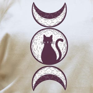 以下が含まれることがあります： 紫色の猫のデザインが円の中に描かれた、三日月が両側に配置されたクリーム色のTシャツ。 Tシャツは丸首で半袖です。「Seasonally Unwell」の文字が丸いバッジにプリントされています。