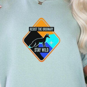 May include: Pale blue t-shirt with a graphic design. The design includes a black fox silhouette, a blue star, and the words "RESIST THE ORDINARY STAY WILD." The graphic is set in a diamond shape with orange, black, and blue. A casual wear item.