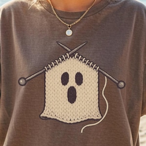 May include: Brown t-shirt with a knitted ghost design. The ghost is beige with black eyes and mouth, and is being knitted with silver needles. The shirt is worn on a beach with a drink in hand.