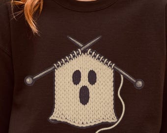 Può includere: Una felpa nera con un disegno di fantasma lavorato a maglia e ferri da maglia. Il fantasma ha tre fori per gli occhi e la bocca spalancata. Il testo "Seasonally Unwell" è stampato in basso a destra nell'immagine.