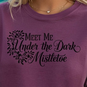May include: A plum-colored t-shirt with the text "Meet Me Under the Dark Mistletoe" in a decorative font. The shirt is worn by someone holding a margarita. The image has a beach theme.