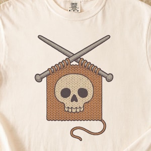 Puede incluir: Camiseta color crema con un diseño de calavera dentro de un cuadrado tejido, con agujas de tejer cruzadas. El diseño es en tonos marrón y crema. La camiseta tiene cuello redondo y mangas enrolladas.