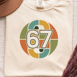 May include: Cream-colored t-shirt with a retro design featuring the number 67 in a circular pattern with orange, green, and teal segments. A brown hat and dried flowers are also in the image. The shirt has the text "Seasonally Unwell".