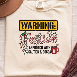May include: A cream-coloured t-shirt featuring the text "WARNING: Festive Approach with Caution & Cocoa." The design includes candy canes, lights, and a mug. A brown hat and dried flowers are also in the image.