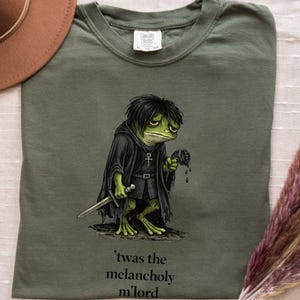 Pode incluir: Camiseta verde oliva com um sapo de desenho animado vestido com uma túnica preta, segurando uma faca e uma cabeça decepada. O texto "'twas the melancholy m'lord" está impresso abaixo. Um chapéu marrom e flores secas também estão na imagem.