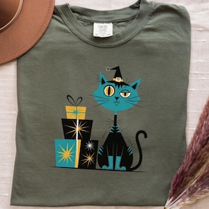 May include: Olive green t-shirt featuring a teal cat wearing a witch hat, with a stack of gift boxes. The cat has one eye closed and is sitting next to the gifts. A brown hat and dried flowers are also in the image.