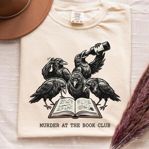 May include: Cream-colored t-shirt featuring a graphic of five black crows gathered around an open book. One crow holds a bottle, and the text "MURDER AT THE BOOK CLUB" is below. A brown hat and dried flowers are also in the image.