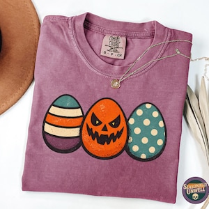 May include: A purple t-shirt with three illustrated eggs. The central egg has a jack-o'-lantern face, with an egg with orange stripes and another with teal polka dots. A brown hat and necklace are visible.