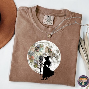 May include: A brown t-shirt with a round neck and short sleeves. The shirt features a graphic of a witch silhouette holding a staff in front of a floral moon. A silver necklace and a brown hat are also visible.