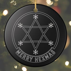 May include: Black round Christmas ornament with a white pentagram design, snowflakes, and the text "MERRY HEXMAS". The ornament is hanging by a gold string against a blurred background of Christmas lights.