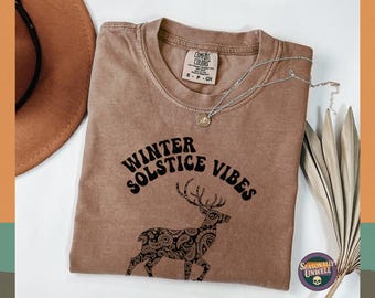 Puede incluir: Una camiseta marrón Comfort Colors con el texto "Winter Solstice Vibes" y un gráfico de ciervo. También se ven un collar plateado con un colgante y un sombrero marrón. La camiseta es talla S-P-CH.