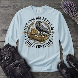 May include: A light blue long-sleeve shirt featuring a graphic of a black raven perched on a book with pocket watches, keys, and gems. The text reads, "MAY YOUR DAY BE FULL OF SHINY TREASURES" in a circular arrangement around the central illustration.