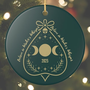 Może przedstawiać: Okrągła, ciemnozielona ozdoba choinkowa ze złotym sznurkiem do zawieszenia. Ozdoba ma złoty wzór z napisem "Believe in Winter's Whisper" i rokiem 2025. Projekt zawiera księżyce, gwiazdy i dzwonek.