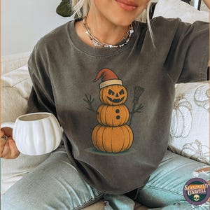 May include: Dark gray t-shirt featuring a Halloween-themed snowman made of pumpkins, wearing a Santa hat and holding a broom. The shirt has a vintage look. A white pumpkin-shaped mug is held in the hand. The text "Seasonally Unwell" is at the bottom.