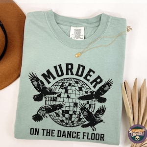 May include: A light green t-shirt with the words "MURDER ON THE DANCE FLOOR" in black, featuring four crows around a disco ball graphic. A gold necklace and a brown hat are also visible.