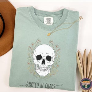 Könnte beinhalten: Hellgrünes T-Shirt mit einem Totenkopf-Motiv, umgeben von einem floralen Kranz. Der Text "Rooted in Chaos" steht darunter. Eine goldene Halskette und ein brauner Hut sind im Hintergrund zu sehen.