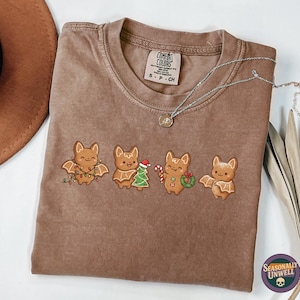 May include: Brown Comfort Colors t-shirt with a crew neck, featuring four embroidered bat characters in festive holiday attire. A silver necklace with a pendant rests on the shirt. The shirt is next to a brown hat and a dried plant.