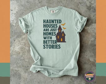 May include: A light green t-shirt with the text "Haunted Houses Are Just Homes With Better Stories" in dark green. The design includes a haunted house and bats in orange. The shirt is folded on a gray surface.
