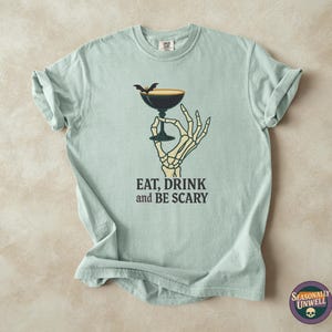 May include: Light blue t-shirt with a graphic of a skeleton hand holding a cocktail glass with a bat on top. The text "EAT, DRINK and BE SCARY" is printed below the graphic. A round logo with a skull is in the bottom right corner.